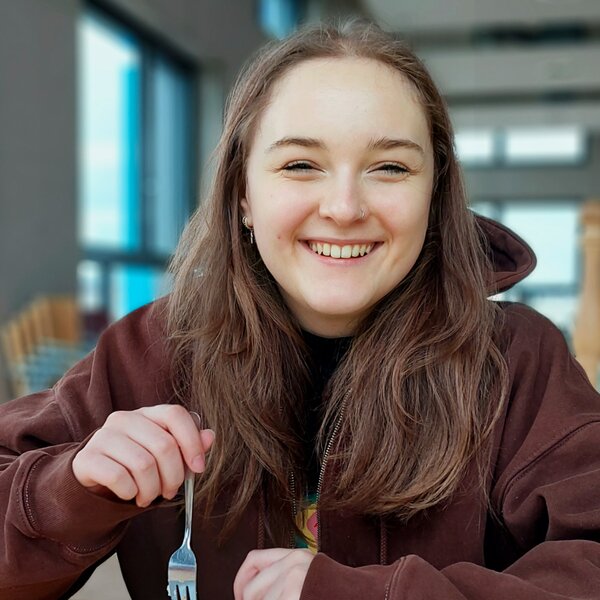 Smiling young woman with brown hair enjoying a meal indoors, with natural light from windows. | © Krankikom GmbH, Photo: Camilla Stengard