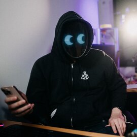 A man wearing a mask sits at a desk and holds a mobile phone in his hand