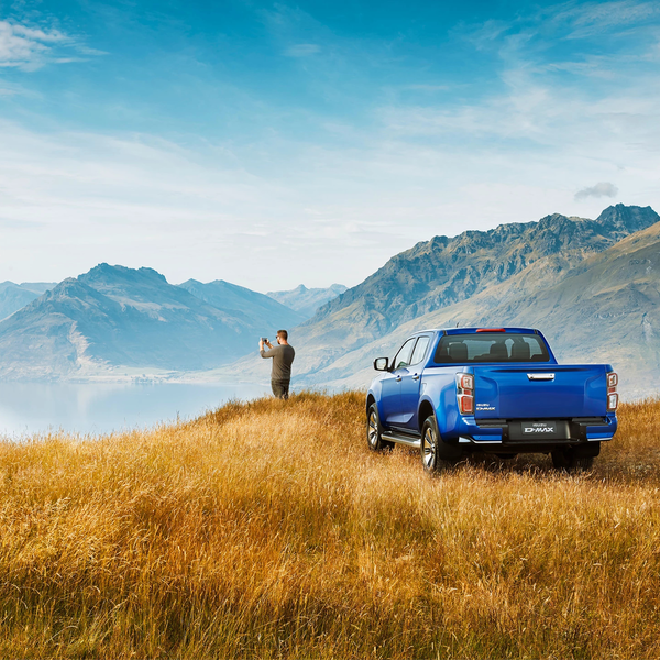 Blue ISUZU D-MAX pickup parked on a hill with a scenic mountain range in the background and a person standing beside | © ISUZU