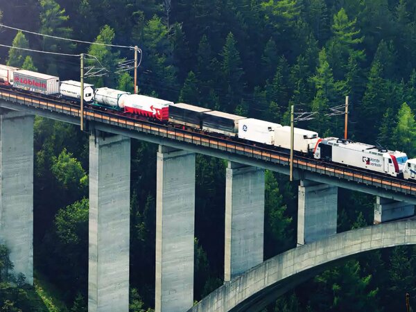 Cargo train traversing a tall bridge over a lush green valley with dense forests.