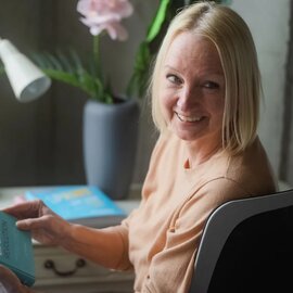 Lächelnde blonde Frau liest ein Buch an einem Schreibtisch mit einer dekorativen Orchidee und Lampe im Hintergrund.