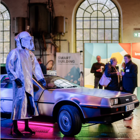 A window mannequin in a futuristic costume standing next to a classic car at a tech event with attendees in the background.
