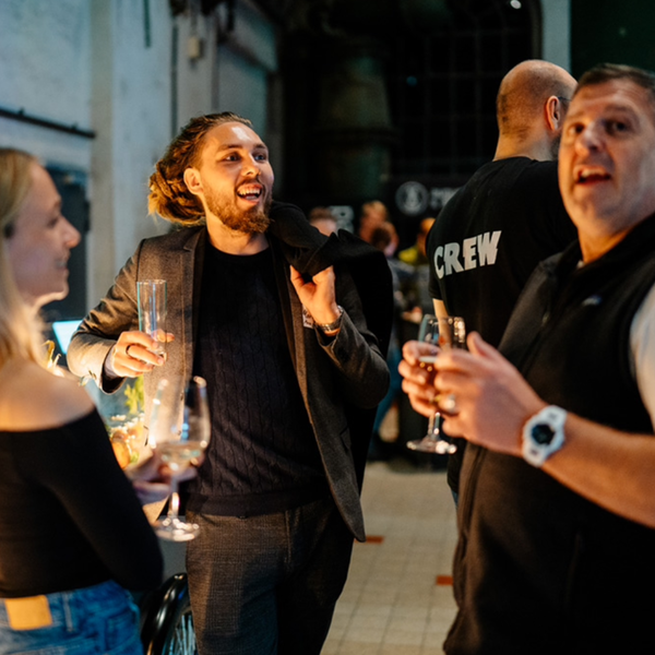 Gruppe von Personen unterhält sich bei einer Veranstaltung mit Getränken in der Hand, darunter ein Mann im Gespräch und ein Crew-Mitglied im Hintergrund.