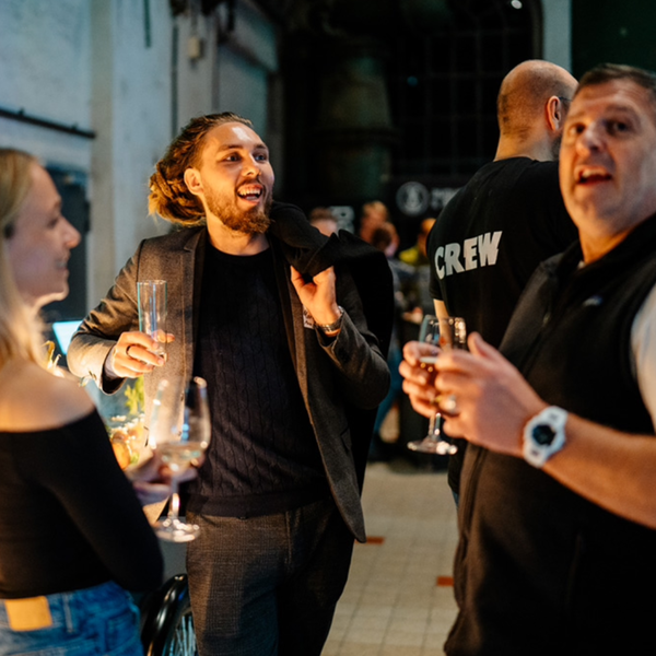 Group of people socializing at an event with drinks in their hands, including a man in conversation and a crew member in the background.