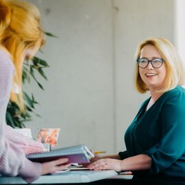 Two professional women engaged in a friendly discussion at a desk with documents and a notebook.