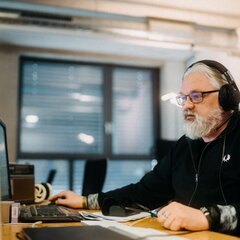 Ein Mann mit Bart und Brille arbeitet in einem Büro an einem Computer und trägt Kopfhörer. | © Photo: Ilja Kagan, 2022