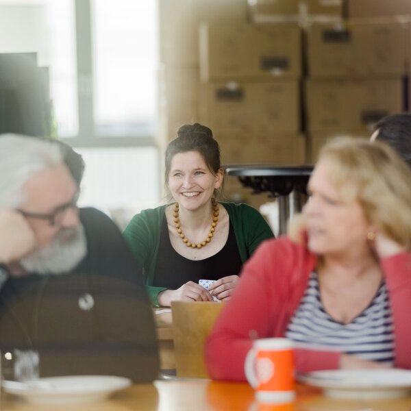 Smiling woman with a wooden necklace sitting at a table in a casual office environment.