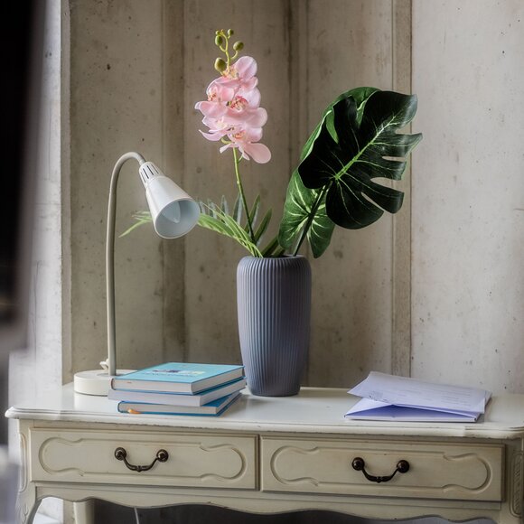 Elegant desk setup with a pink orchid in a vase, a monstera leaf, a lamp, and some books on a vintage table.