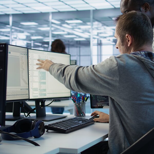 Mann zeigt mit dem Finger auf einen Monitor mit Code oder Tabellen, daneben Tastatur und Headset, im Hintergrund weitere Personen im Büro.