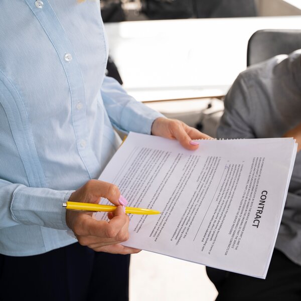 Nahaufnahme einer Person im hellblauen Hemd, die ein Dokument mit der Aufschrift „Contract" und einem gelben Stift hält.