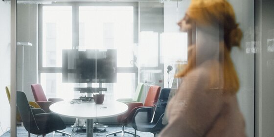 Blurry motion of a person walking past a modern office meeting room with a round table and colorful chairs. | © Photo: Ilja Kagan, 2022