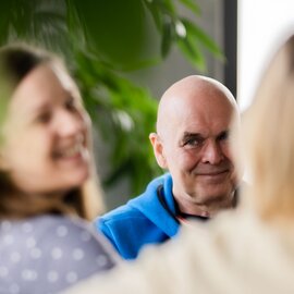 Lächelnder Mann in einem blauen Pullover, der sich mit unscharfen Figuren im Vordergrund unterhält. | © Photo: Ilja Kagan, 2022