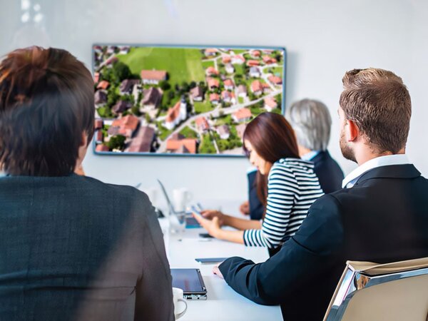 Menschen sitzen gemeinsam an einem Bürotisch und schauen auf einen großen Monitor.