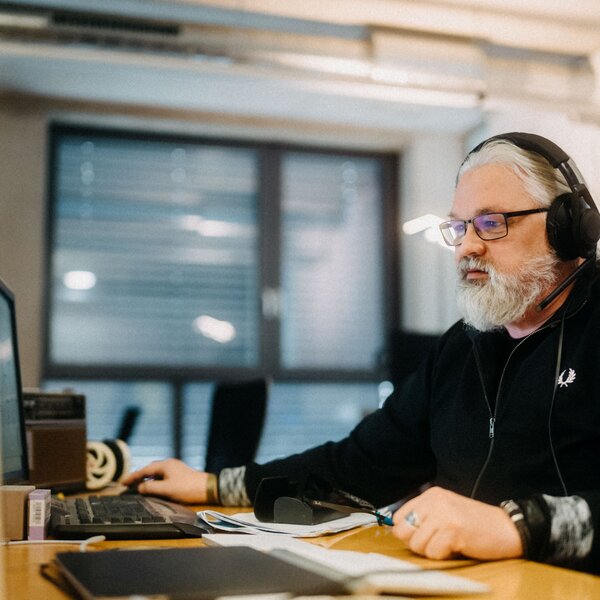 Ein Mann mit Bart und Brille arbeitet in einem Büro an einem Computer und trägt Kopfhörer. | © Photo: Ilja Kagan, 2022