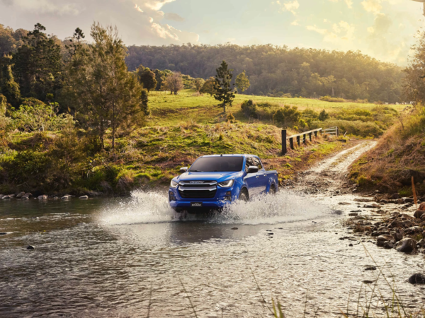 Blue ISUZU D-MAX pickup truck driving through a shallow stream in a lush countryside setting. | © ISUZU