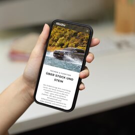 Hand hält ein Smartphone, auf dem Bildschirm ist die Webseite von ISUZU D-MAX zu sehen
