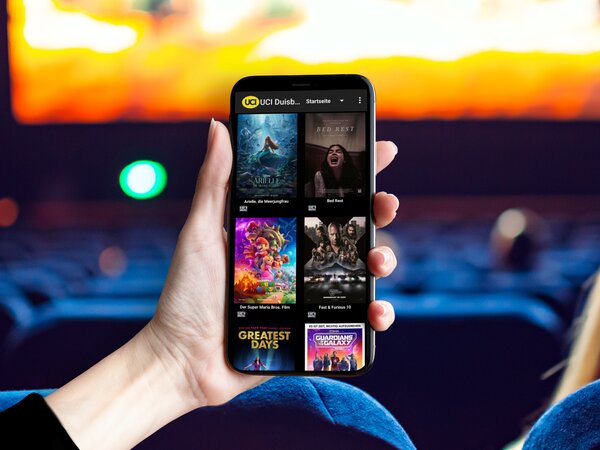 Person hält ein Smartphone mit geöffneter UCI Kinowelt-App in einem Kinosaal.
