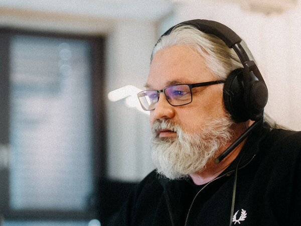 Ein Mann mit Bart und Brille arbeitet in einem Büro an einem Computer und trägt Kopfhörer. | © Photo: Ilja Kagan, 2022