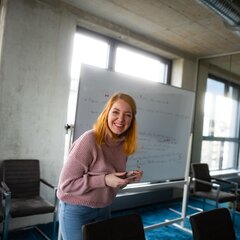 Eine lächelnde Frau steht vor einem Whiteboard in einem modernen Büro. Sie trägt einen rosa Pullover und schaut in die Kamera. | © Photo: Ilja Kagan, 2022