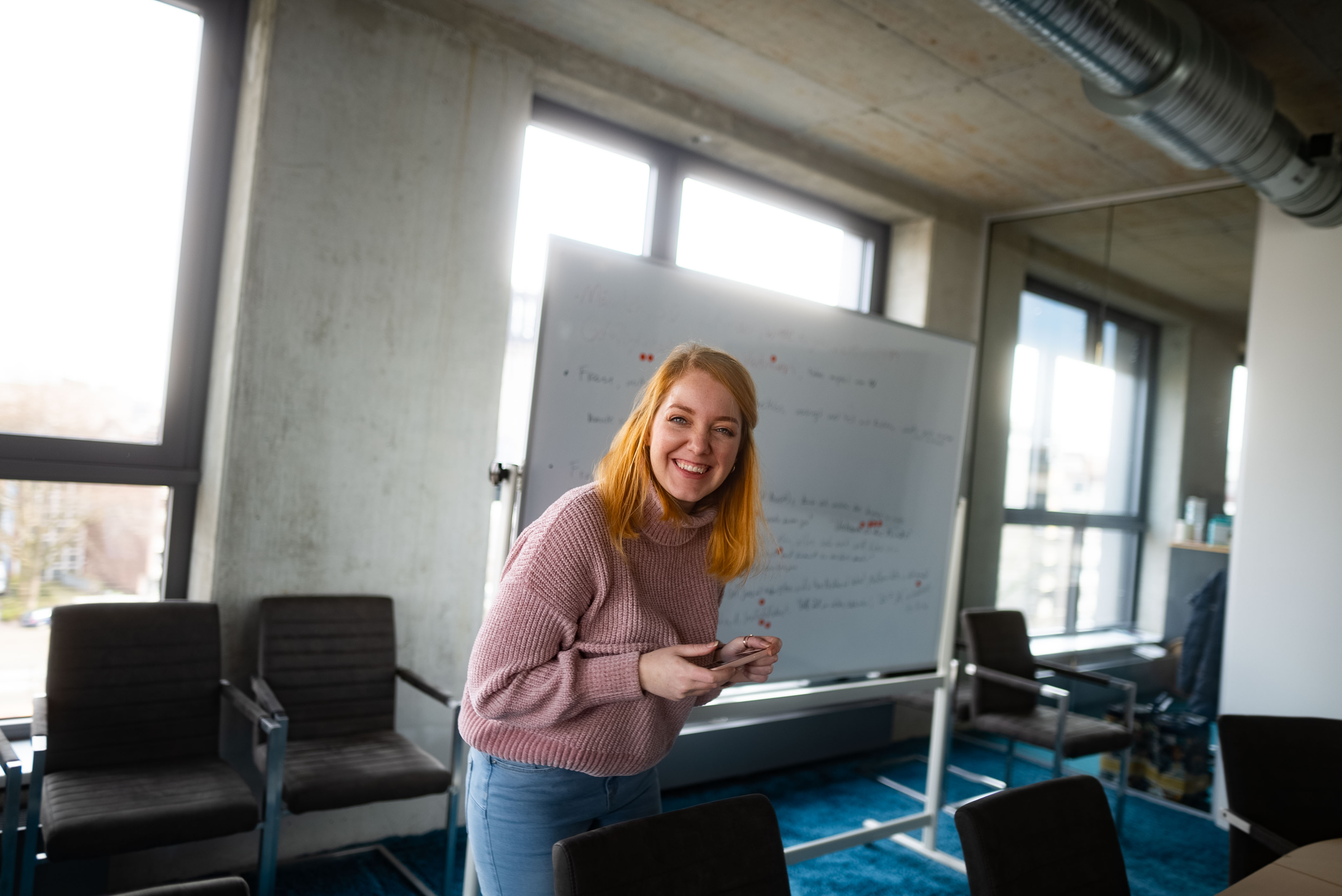 Eine lächelnde Frau steht vor einem Whiteboard in einem modernen Büro. Sie trägt einen rosa Pullover und schaut in die Kamera. | © Photo: Ilja Kagan, 2022