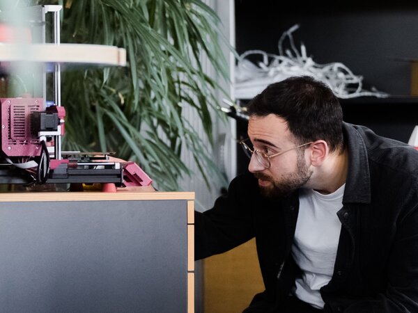 Mann beobachtet einen in Betrieb befindlichen 3D-Drucker in einem kreativen Büroumfeld. | © Photo: Ilja Kagan, 2022