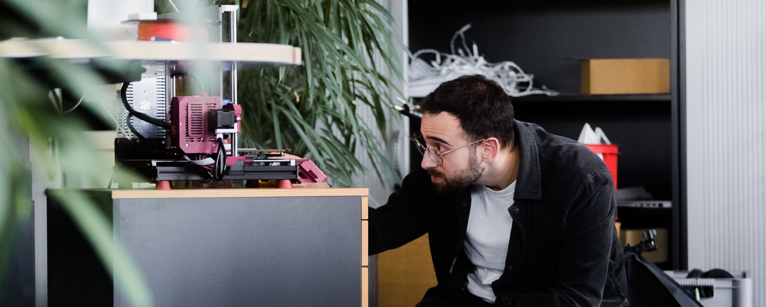 Mann mit Brille kniet vor einem Schreibtisch und überprüft einen 3D-Drucker im Büro. | © Photo: Ilja Kagan, 2022