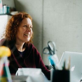 Fröhliche Frau mit lockigen roten Haaren lächelt beim Arbeiten an ihrem Laptop in einem modernen Büroumfeld.